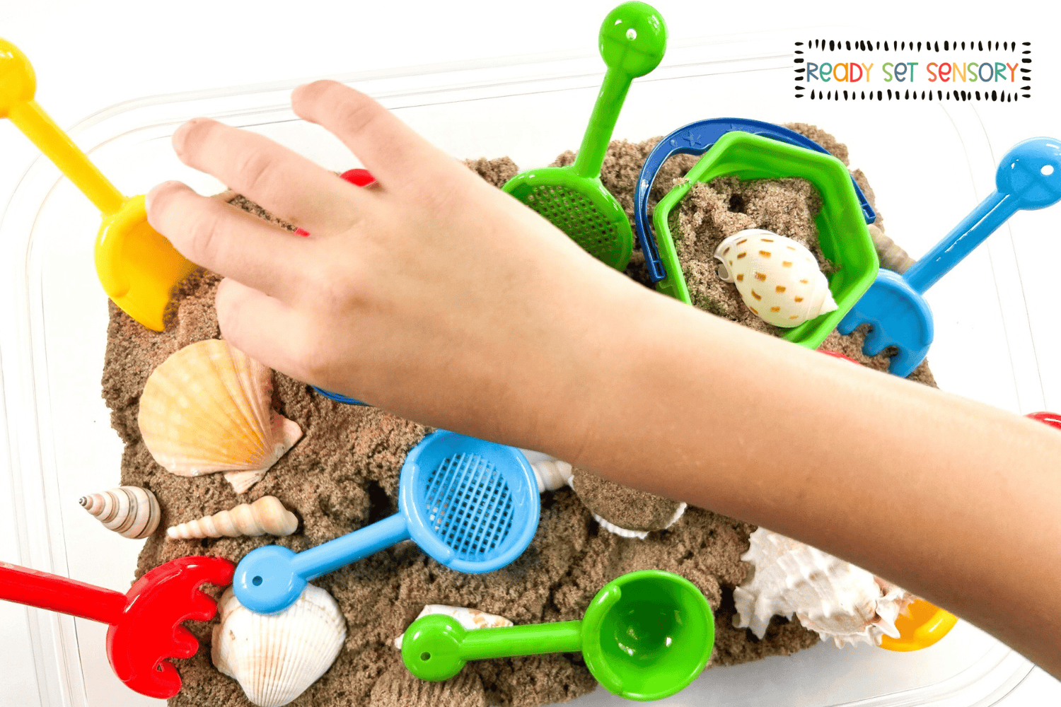 Toddler playing with beach sensory bin using sand tools