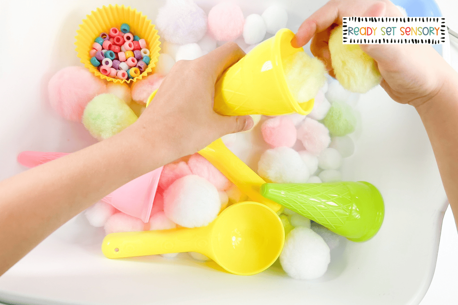 Child scooping pom poms in an ice cream sensory bin with toy cones