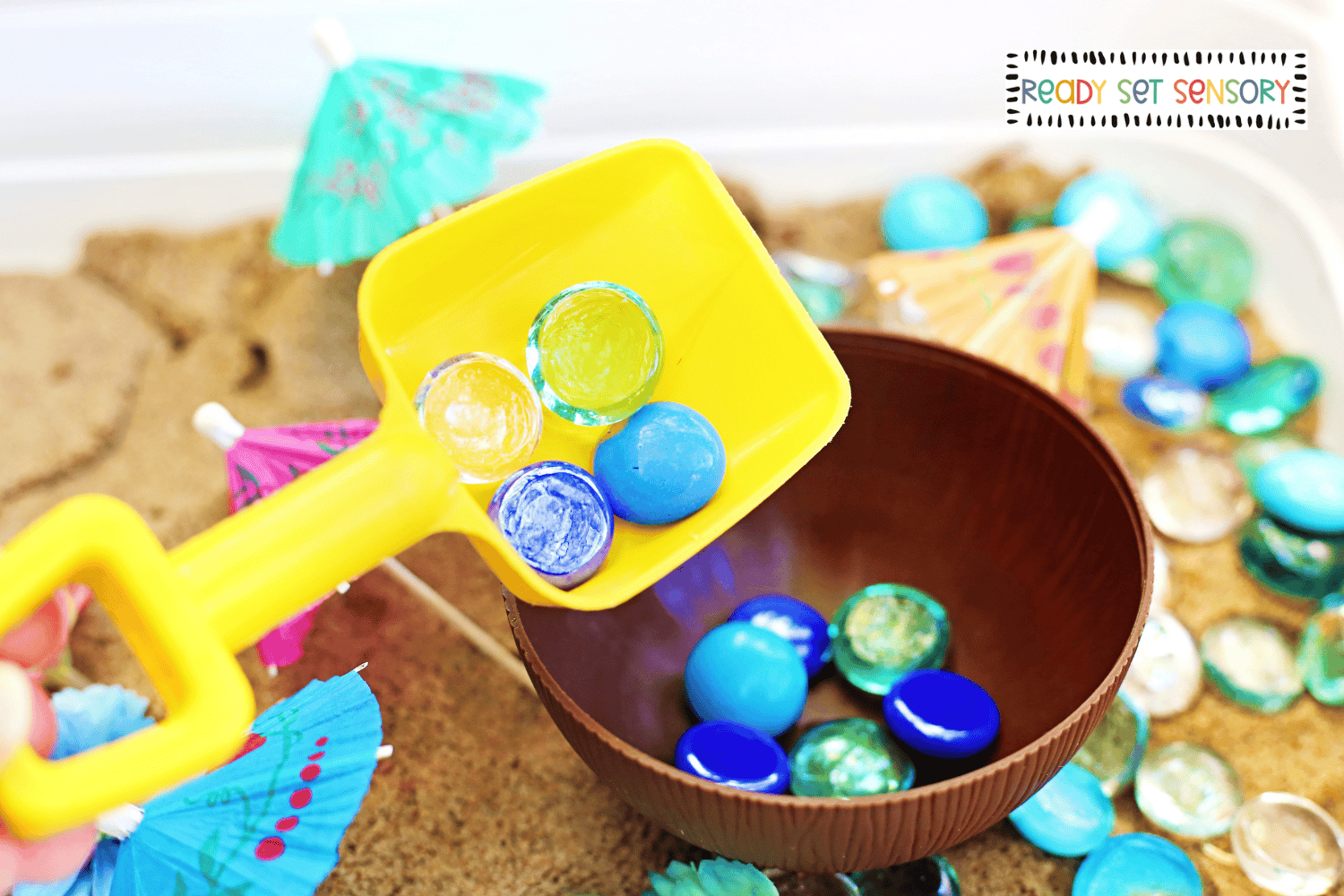 Scooping colorful gems in tropical sensory bin with sand