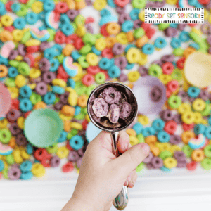Child holding scoop with purple cereal in rainbow color sorting sensory bin